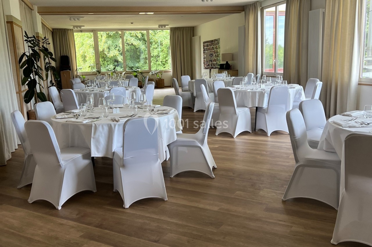 Salle lumineuse avec tables rondes dressées, nappes blanches et chaises couvertes, prête pour un événement.