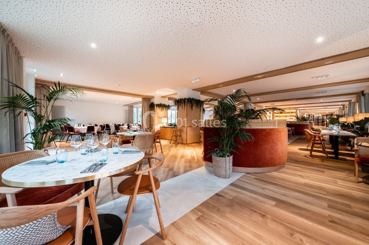 Salle de restaurant lumineuse avec tables en bois, chaises modernes, plantes décoratives et bar en arrière-plan.