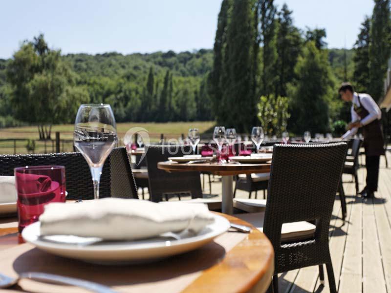 Terrasse d'un restaurant avec des tables dressées, vue sur un paysage verdoyant et un serveur en arrière-plan.