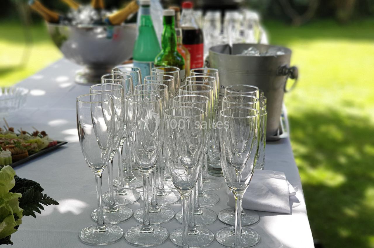 Verres à champagne alignés sur une table blanche avec des boissons et un seau à glace en arrière-plan, en extérieur.