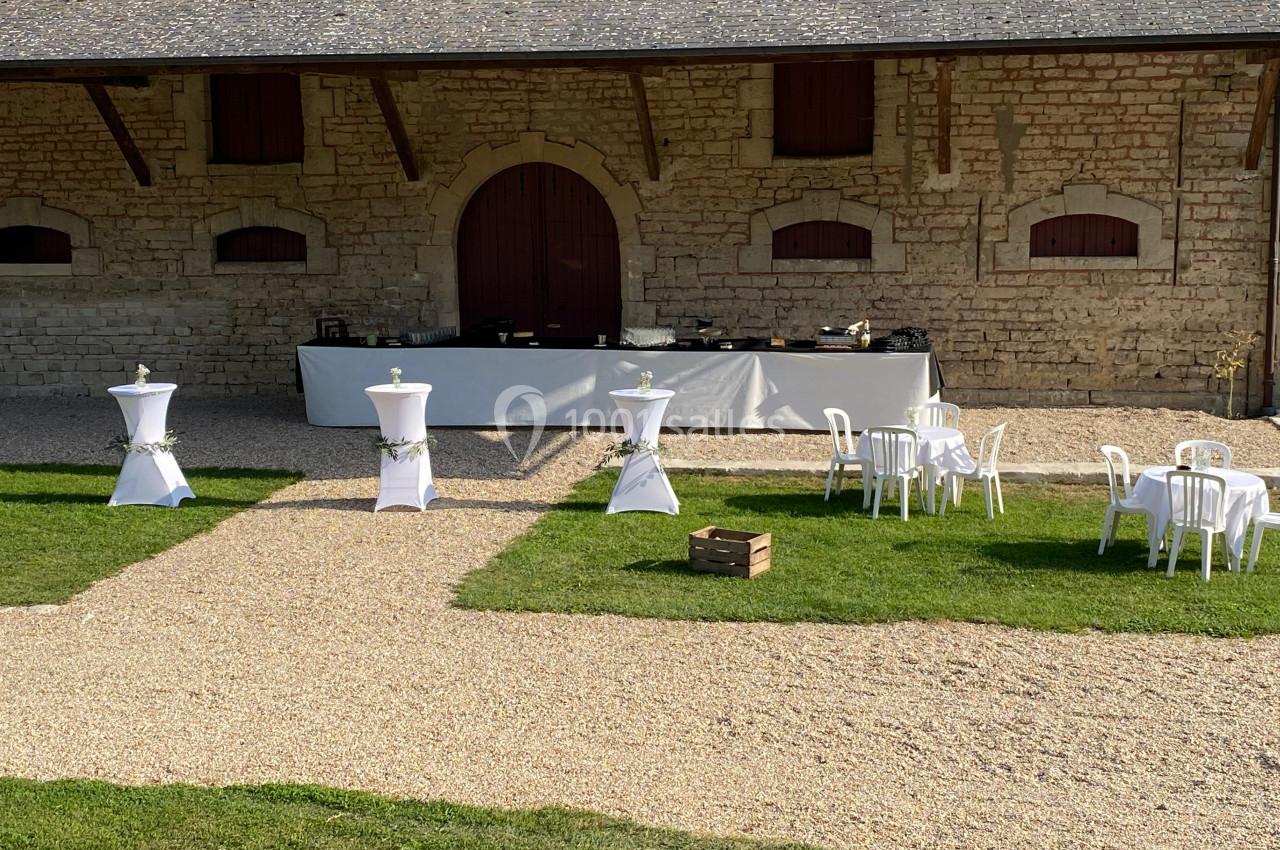 Espace extérieur avec tables hautes et basses dressées devant un bâtiment en pierre, pour un événement en plein air.