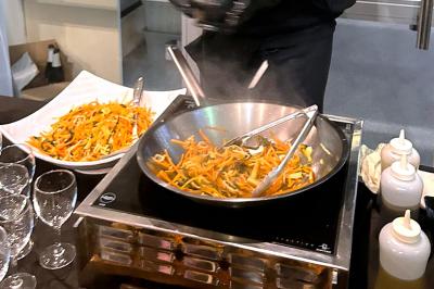 Un chef cuisine des légumes sautés dans une poêle en inox, avec un plat de légumes préparés à côté.