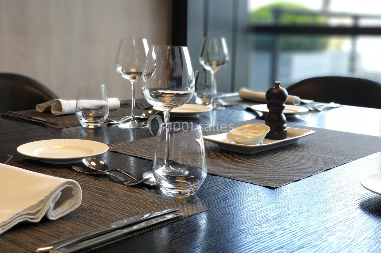 Table dressée avec des verres, assiettes, couverts et serviettes, sur un fond lumineux avec une vue extérieure floue.