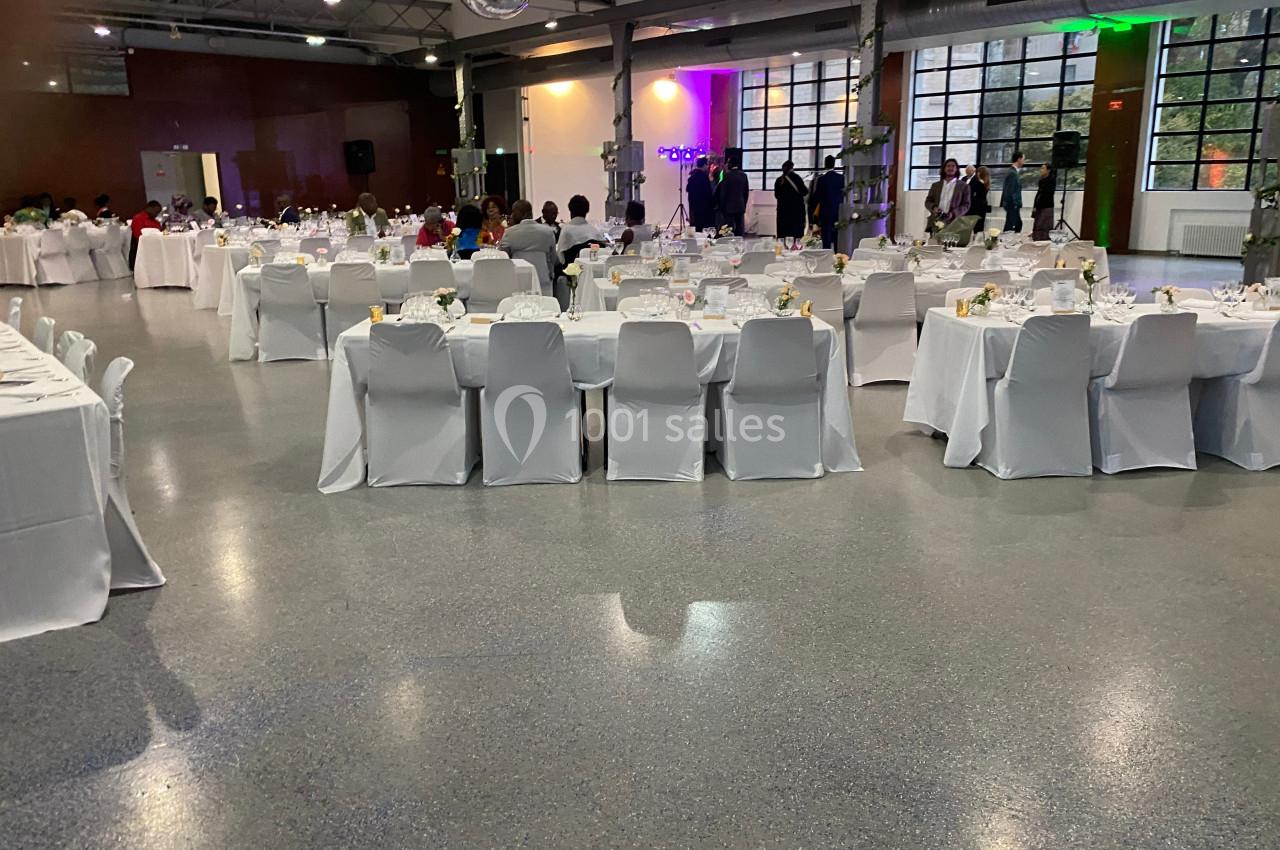 Salle de réception avec tables rondes et rectangulaires dressées, chaises blanches et invités en arrière-plan.