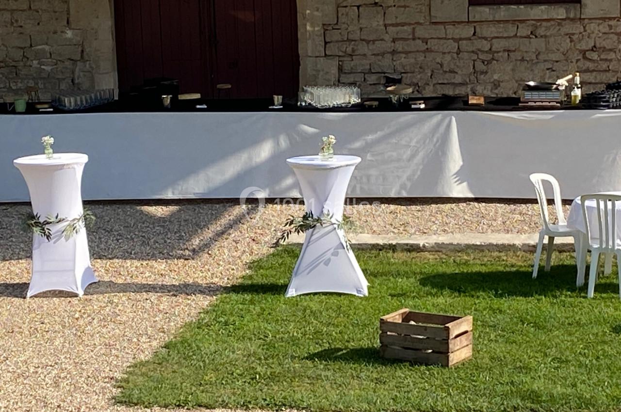 Deux tables hautes décorées de nappes blanches et feuillages, devant un buffet en extérieur sur gravier et pelouse.