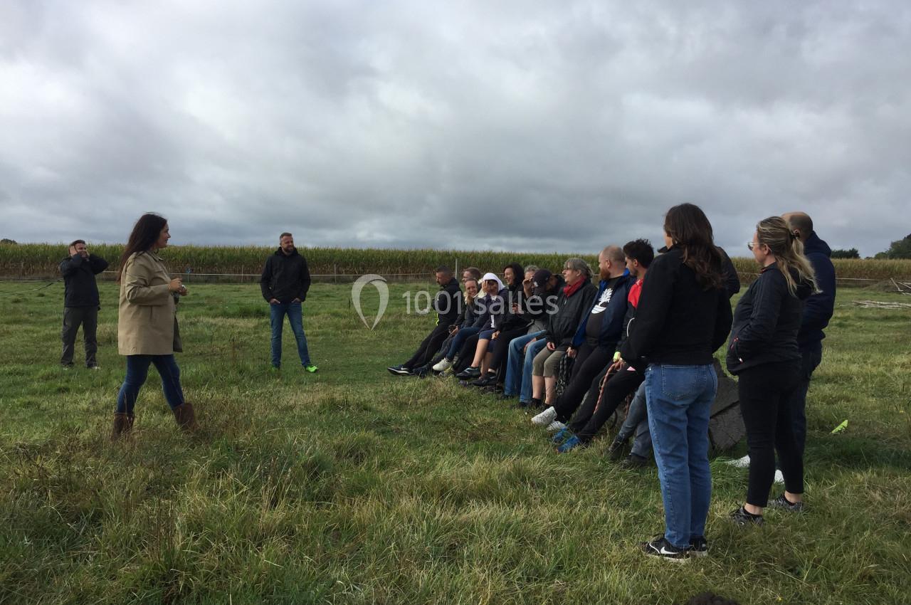 Un groupe de personnes assises en plein air sur une prairie, écoutant deux intervenants debout.
