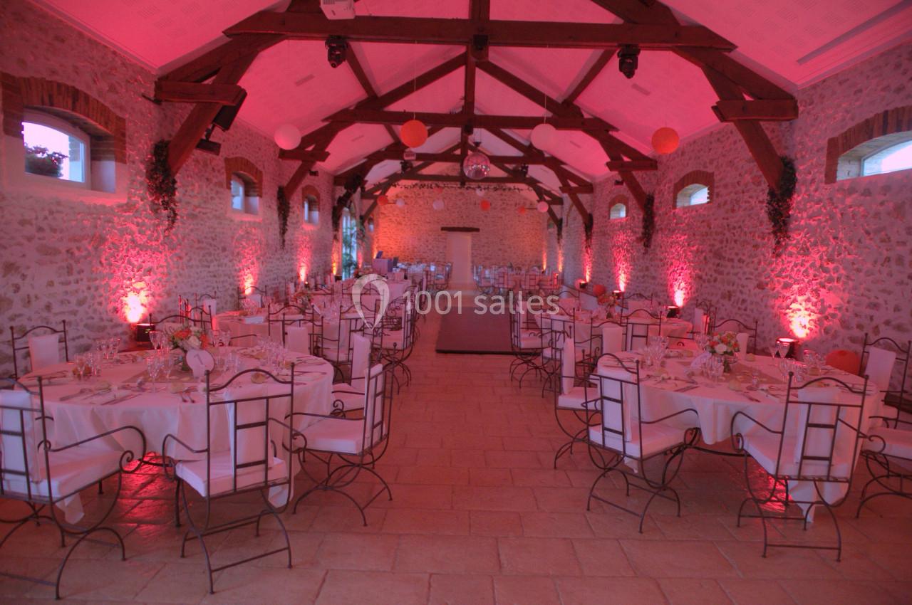 Salle de réception en pierre avec poutres apparentes, tables dressées et éclairage d'ambiance rose.