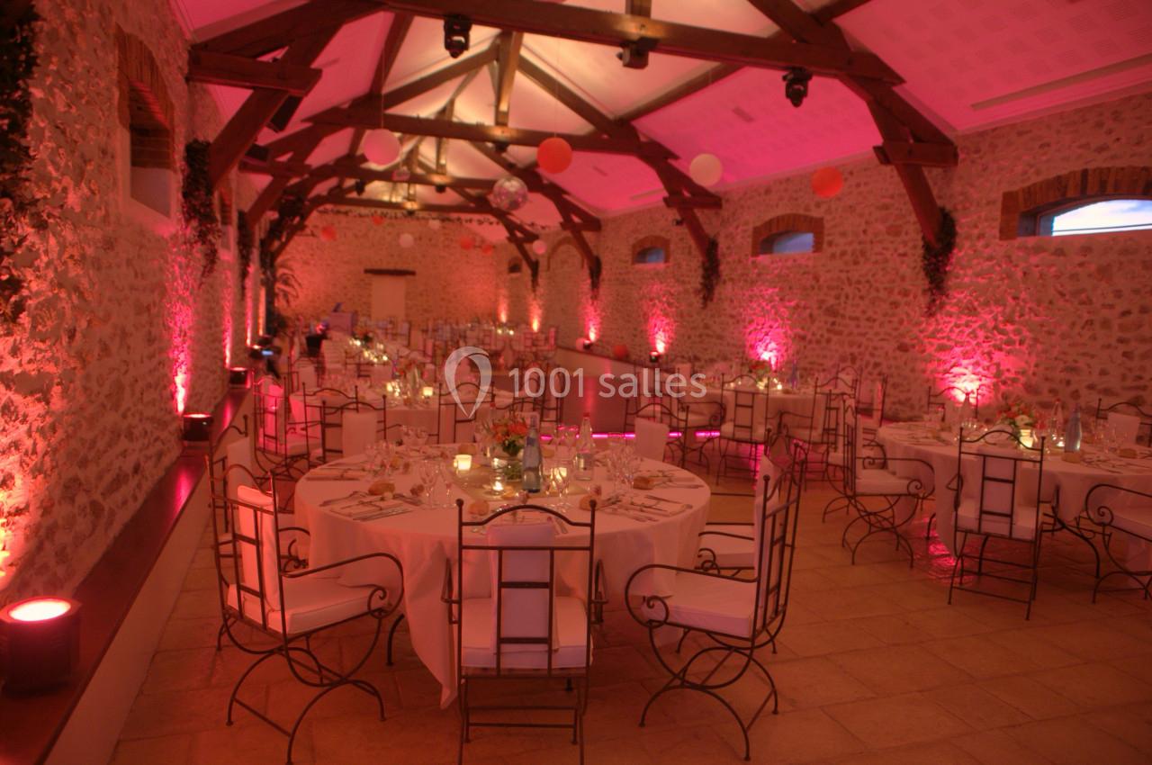 Salle en pierre avec poutres apparentes, tables rondes dressées pour un événement, éclairage rose tamisé.