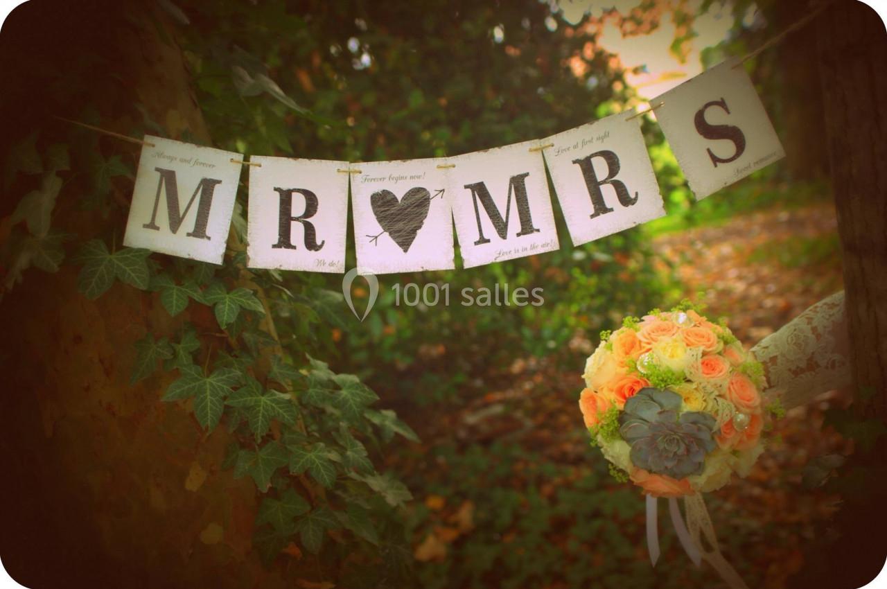 Guirlande ’Mr & Mrs’ suspendue entre des arbres, avec un bouquet de roses pastel en premier plan.