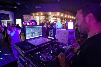 Un DJ mixe de la musique dans un bar animé avec des lumières colorées et des danseurs en arrière-plan.