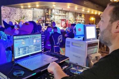 Un DJ mixe de la musique dans un bar animé avec des clients déguisés et des lumières colorées en arrière-plan.