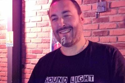 Homme souriant portant un t-shirt noir avec l'inscription ’Sound Light’, devant un mur de briques rouges.
