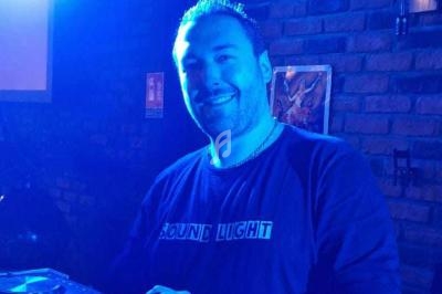 Homme souriant dans une ambiance lumineuse bleutée, debout devant un mur de briques avec un équipement audio visible.