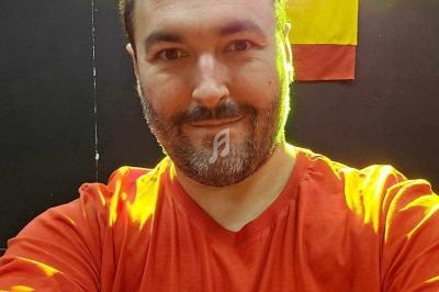 Homme souriant portant un t-shirt rouge, éclairé par des lumières colorées, avec un drapeau en arrière-plan.
