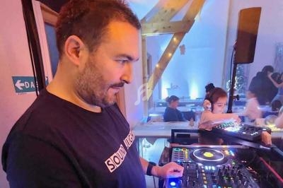 Un DJ mixe de la musique sur une console dans une salle éclairée, avec des personnes en arrière-plan.