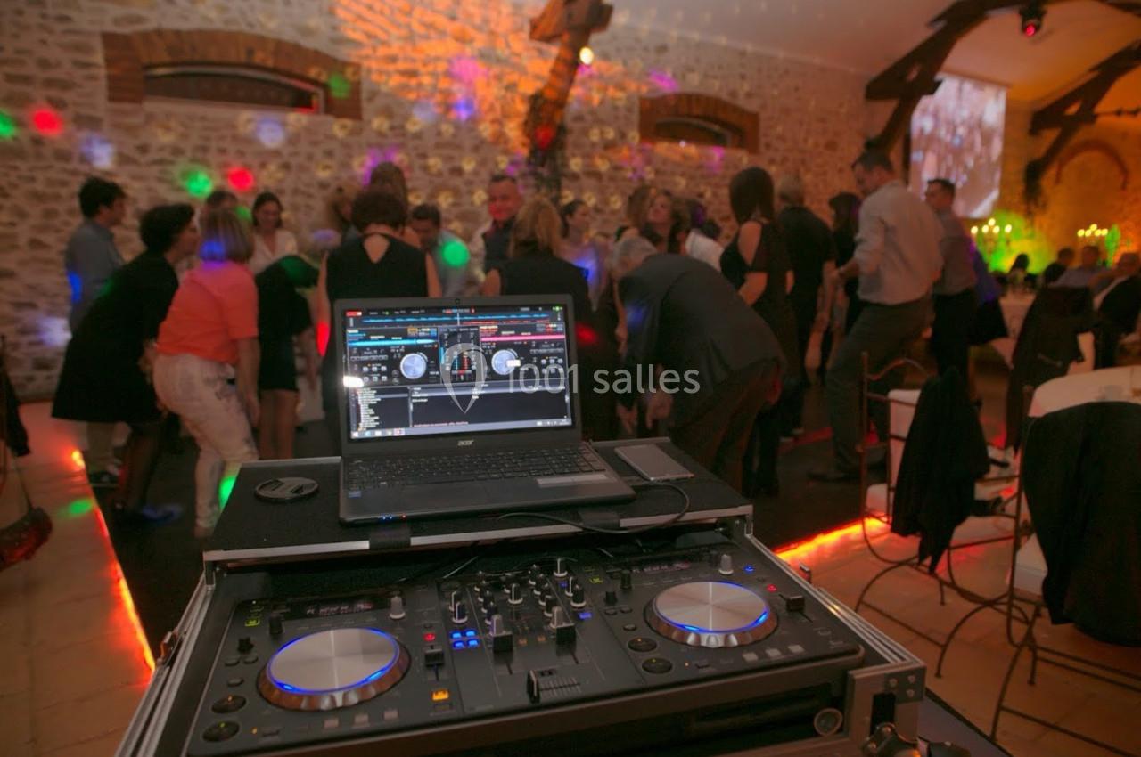 Console de DJ avec ordinateur portable en premier plan, invités dansant sous des lumières colorées dans une salle en pierre.