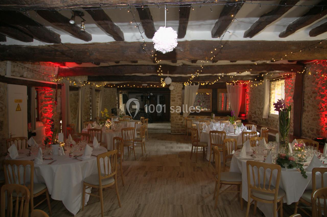 Salle de réception rustique décorée de guirlandes lumineuses, tables rondes dressées avec nappes blanches et chaises en bois.