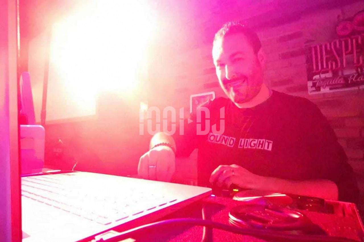 Un homme souriant mixe de la musique sur un équipement DJ dans une ambiance lumineuse rouge.