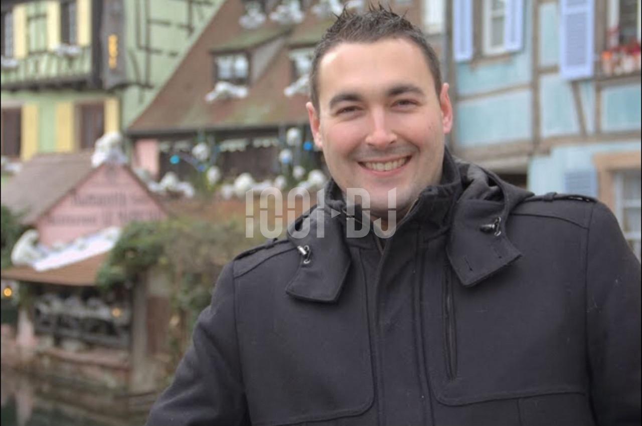 Homme souriant en manteau noir devant des maisons à colombages colorées dans un décor urbain hivernal.