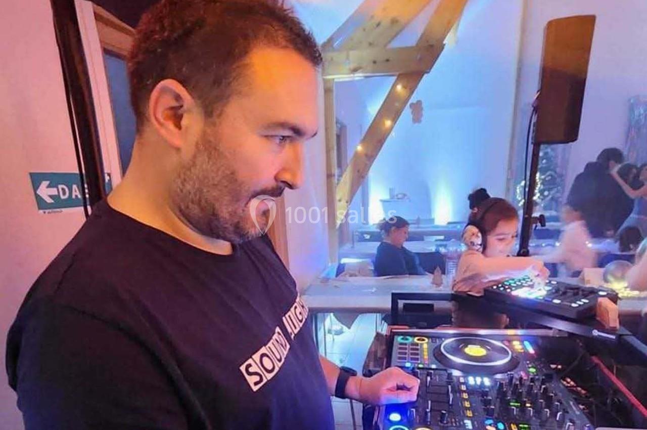 Un DJ mixe sur une console dans une salle éclairée, avec des personnes en arrière-plan.