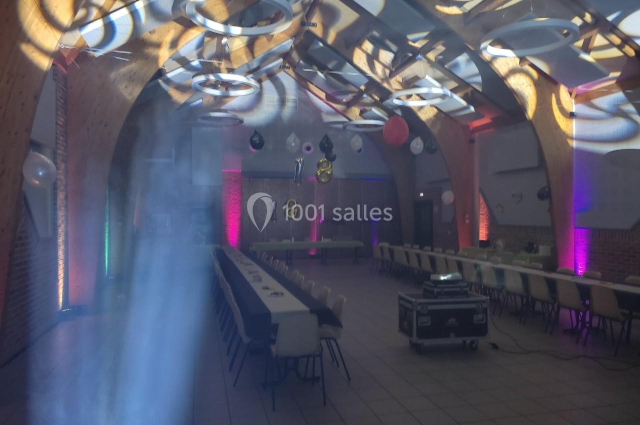 Salle décorée pour un événement, avec tables alignées, éclairages colorés et ballons suspendus au plafond.