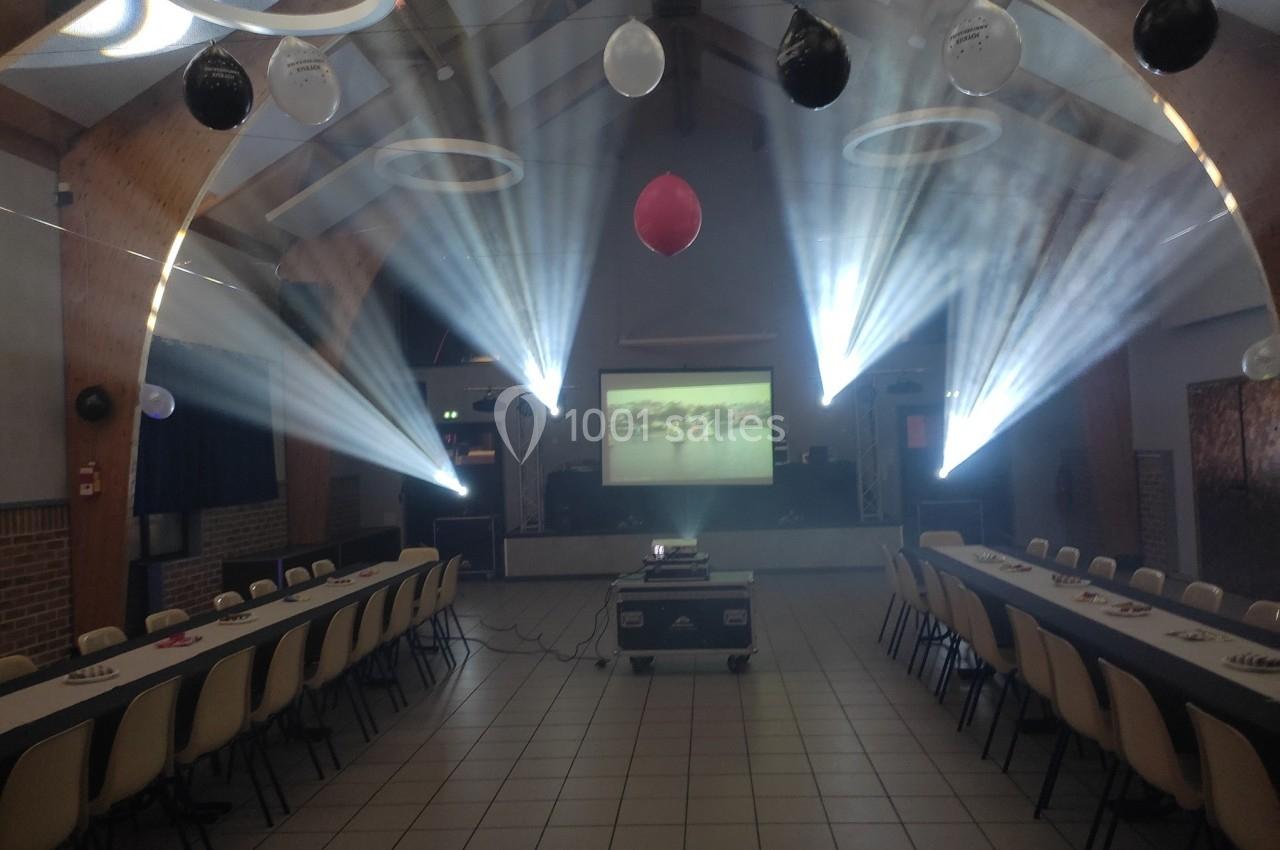 Salle décorée avec des ballons, tables alignées et projecteur diffusant une image sur un écran sous des jeux de lumière.