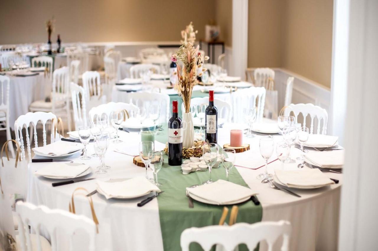Tables décorées pour un événement, avec nappes blanches, chemins de table verts, bouteilles de vin et compositions florales.