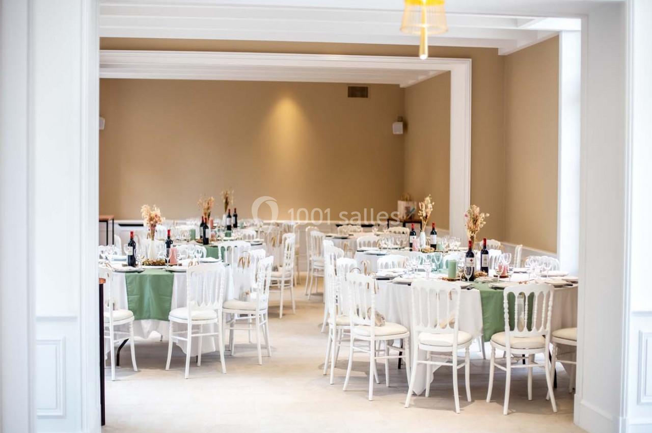 Salle de réception lumineuse avec des tables dressées, nappes vertes et blanches, décorations florales et chaises blanches.