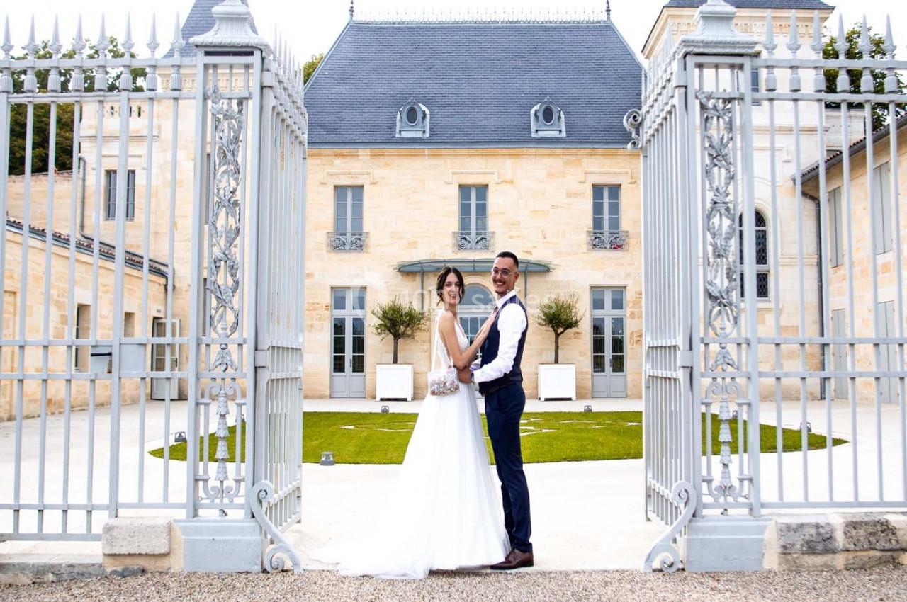 Un couple en tenue de mariage pose devant un portail ouvert menant à une grande bâtisse en pierre.