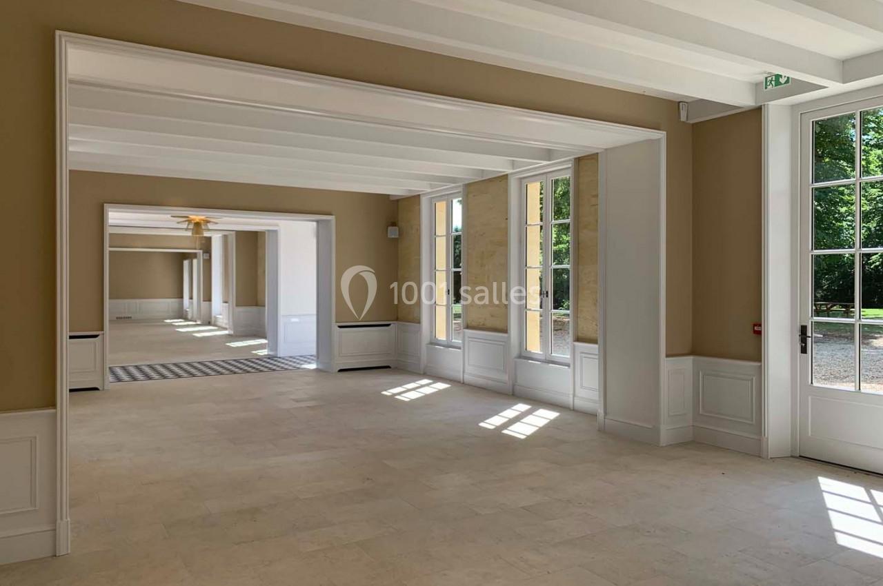 Intérieur lumineux avec grandes fenêtres, murs beige clair, sol en pierre et vue sur un jardin verdoyant.