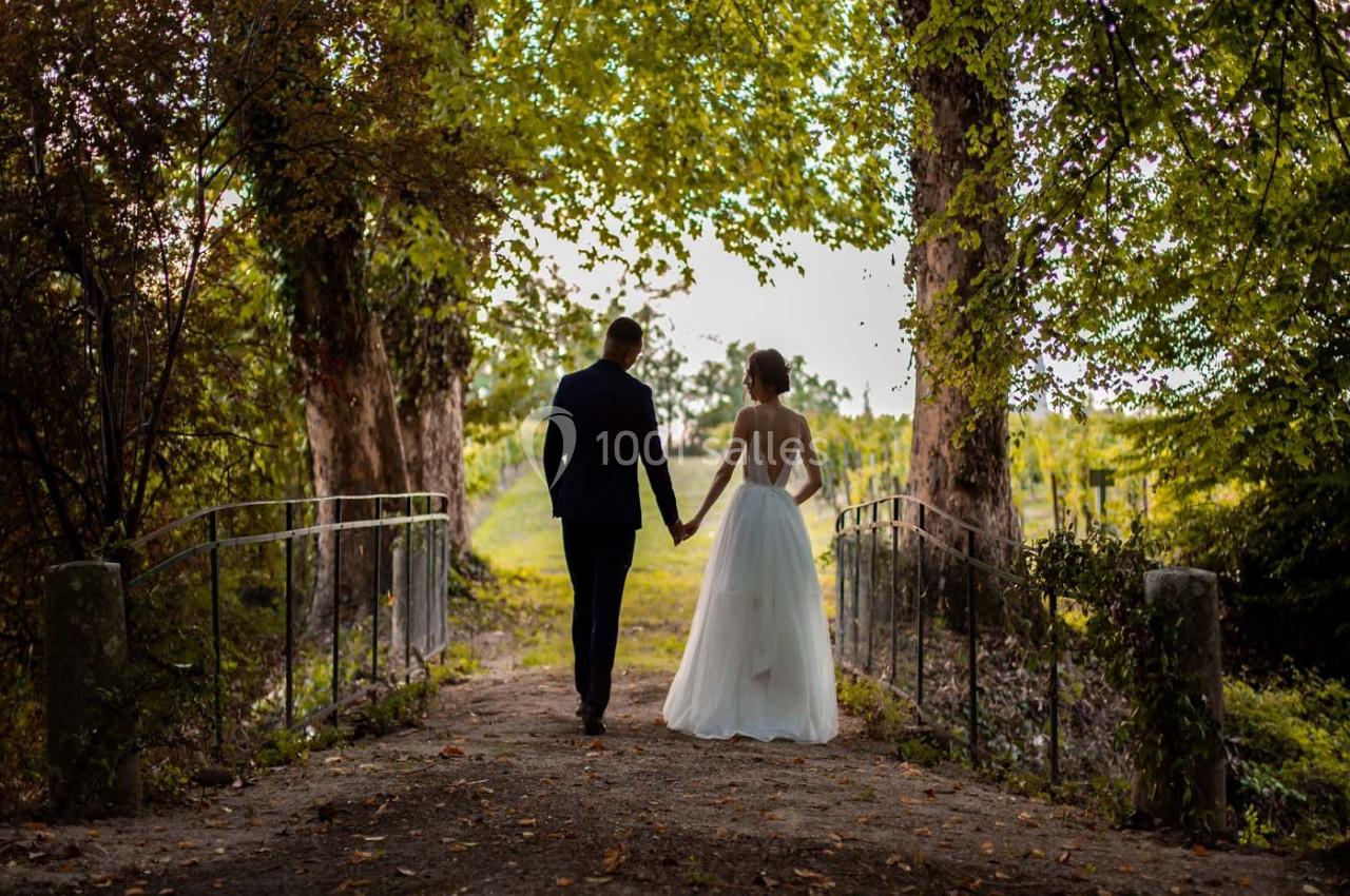 Un couple en tenue de mariage marche main dans la main sur un chemin bordé d'arbres verdoyants.