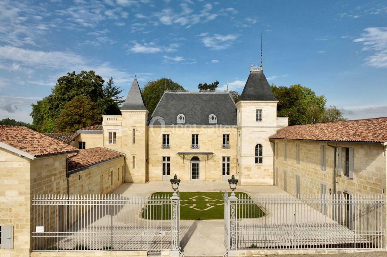 Façade d'un château en pierre entouré de bâtiments annexes, avec une grille et une cour aménagée au premier plan.