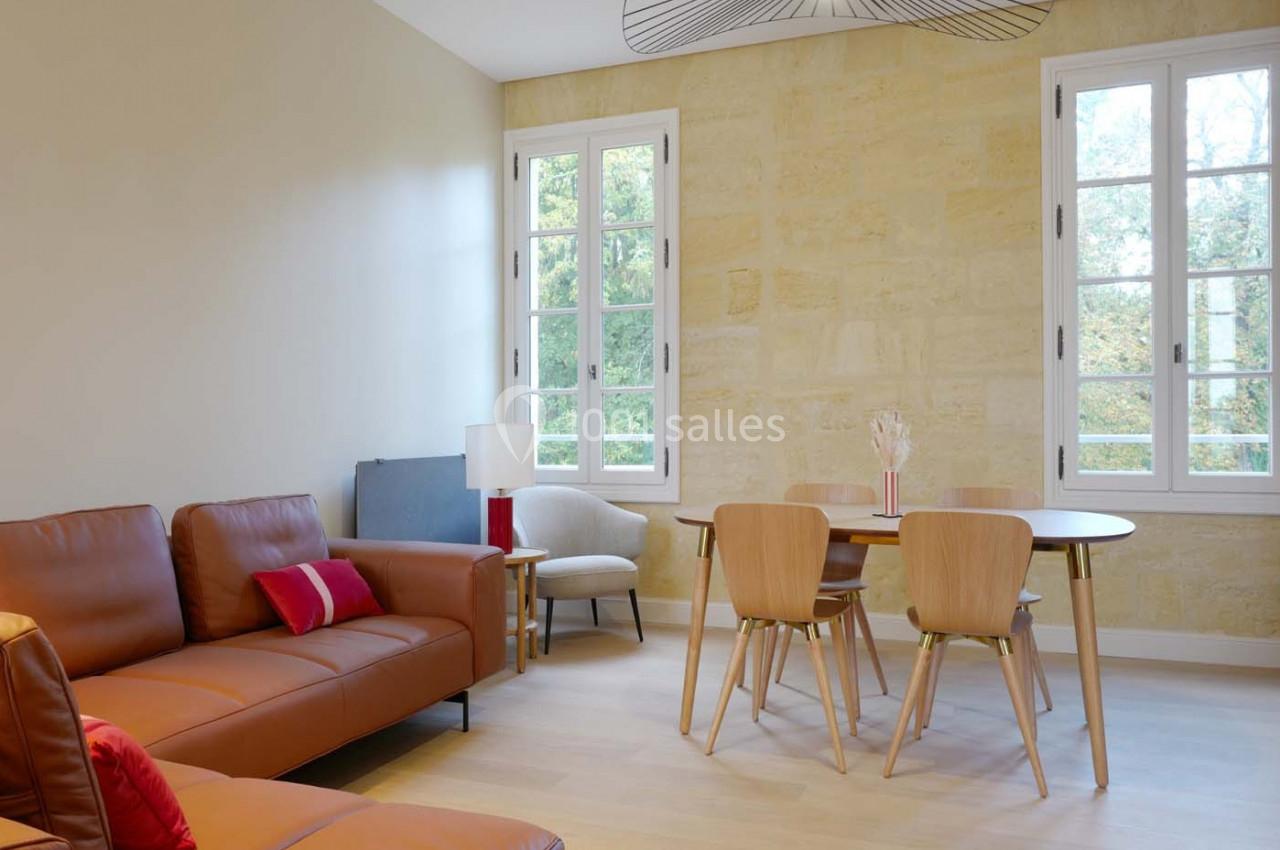 Salon lumineux avec murs en pierre, canapé en cuir, table en bois entourée de chaises et grandes fenêtres.