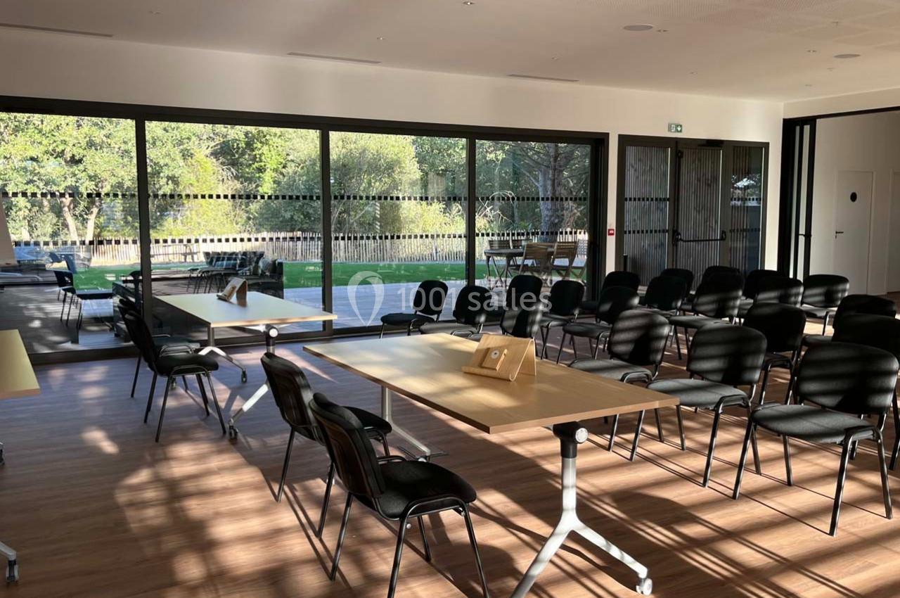 Salle lumineuse avec baies vitrées, aménagée avec des chaises et des tables, donnant sur un espace extérieur verdoyant.