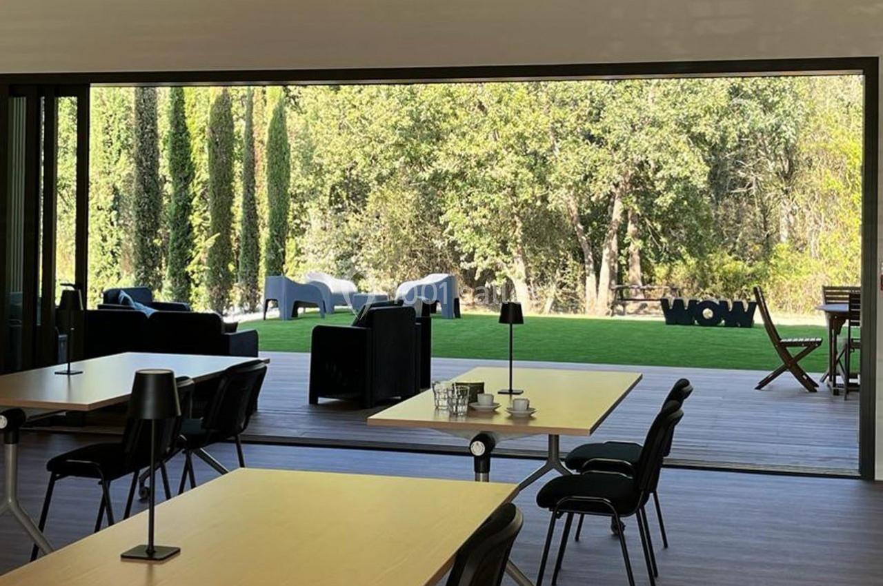 Vue d'une terrasse ouverte sur un jardin arboré, avec des tables, des chaises et des fauteuils extérieurs.