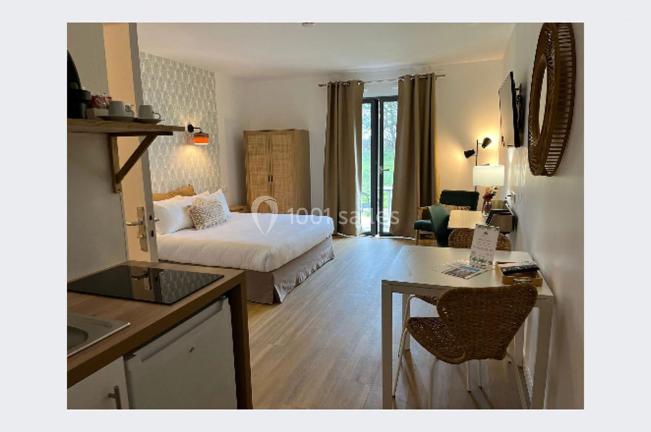 Chambre lumineuse avec lit double, bureau, coin salon, kitchenette et vue sur un jardin à travers une baie vitrée.