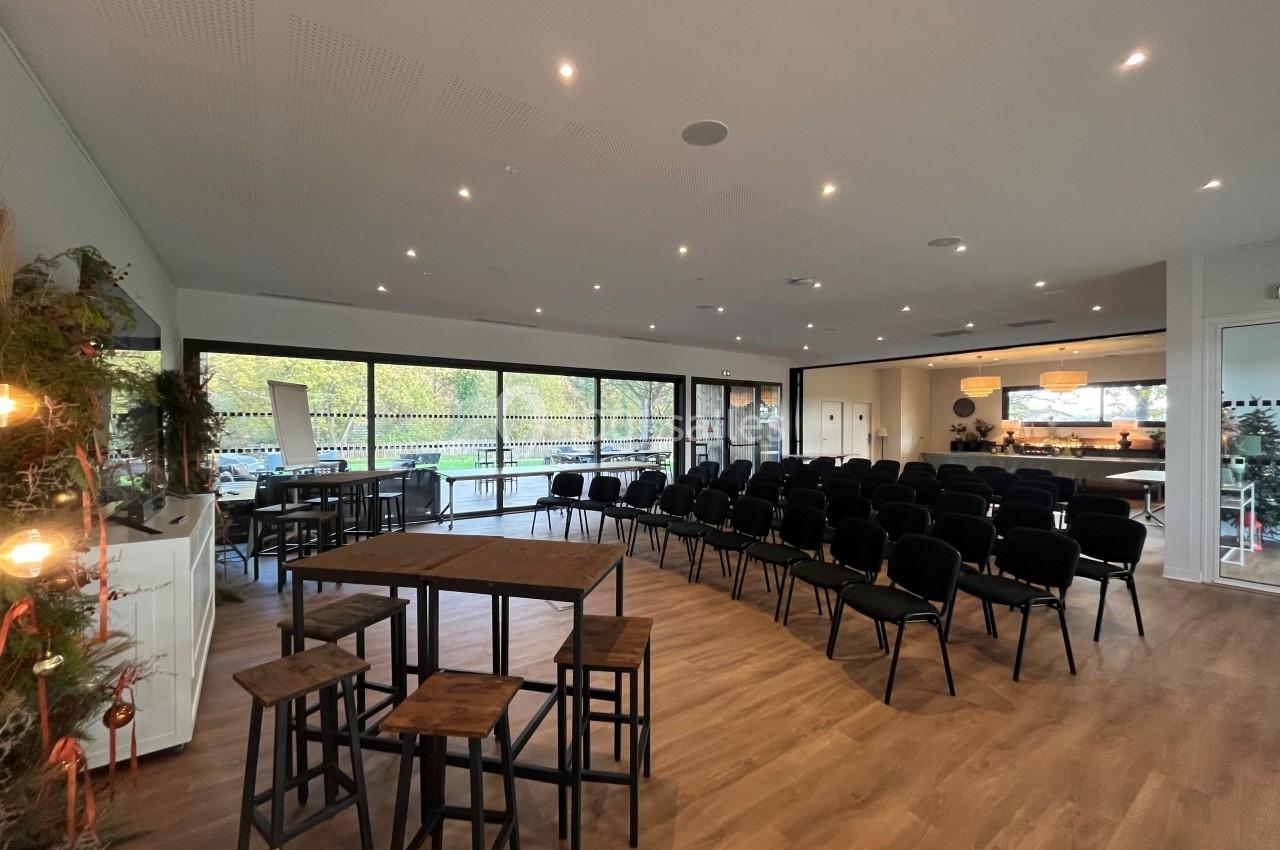 Salle lumineuse avec baies vitrées, chaises alignées pour une présentation et tables hautes en bois au premier plan.