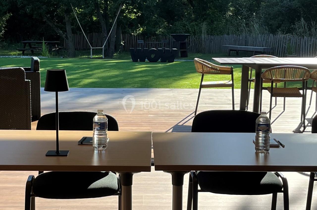 Deux tables avec chaises, lampes et bouteilles d'eau, devant une terrasse ensoleillée donnant sur un jardin verdoyant.