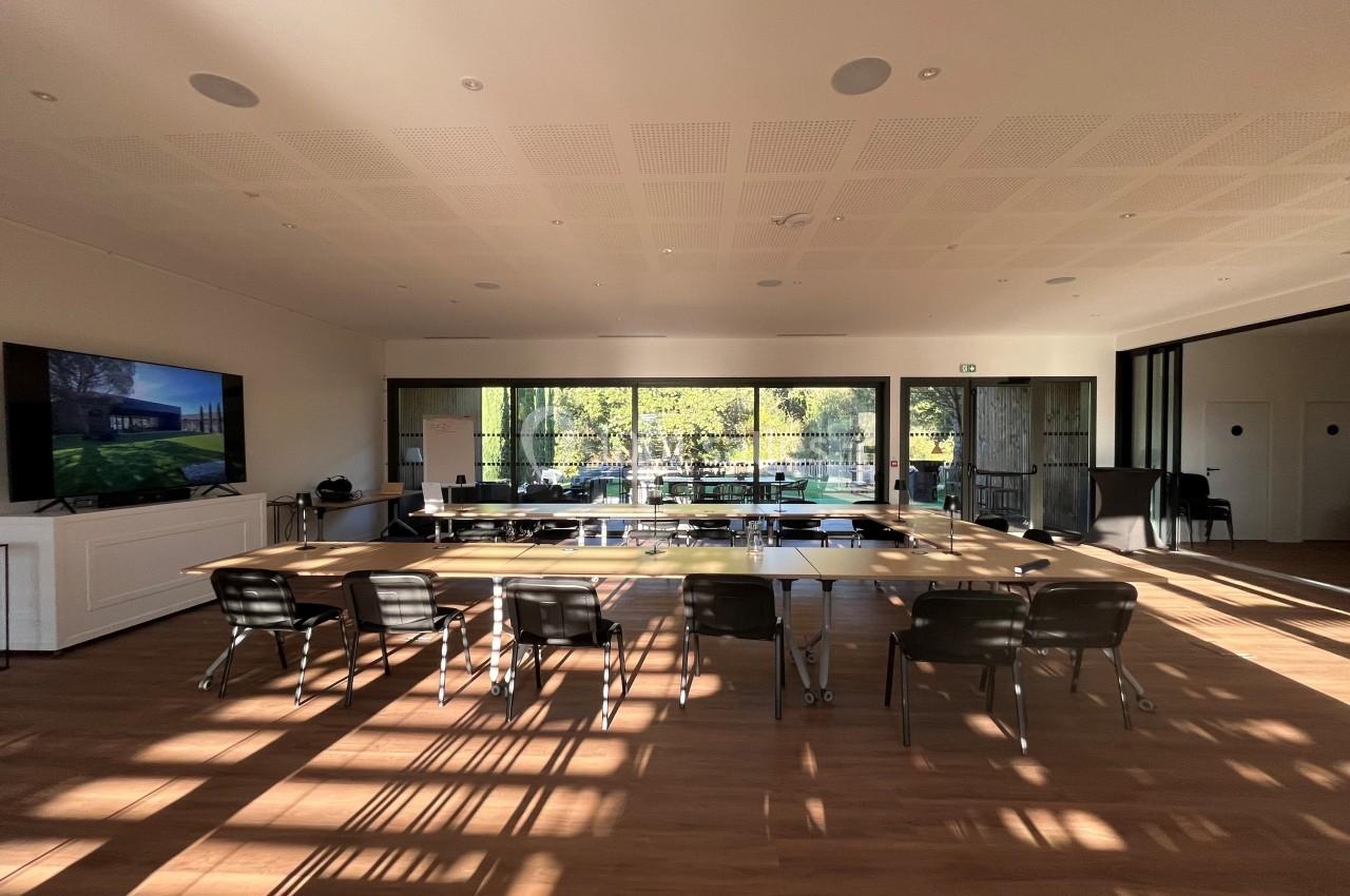 Salle de réunion lumineuse avec une grande table en bois, des chaises noires et une vue sur un jardin à travers des baies…