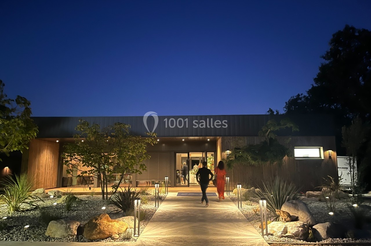 Entrée d'un bâtiment moderne éclairé la nuit, avec un chemin bordé de plantes et de lumières, deux personnes marchant.