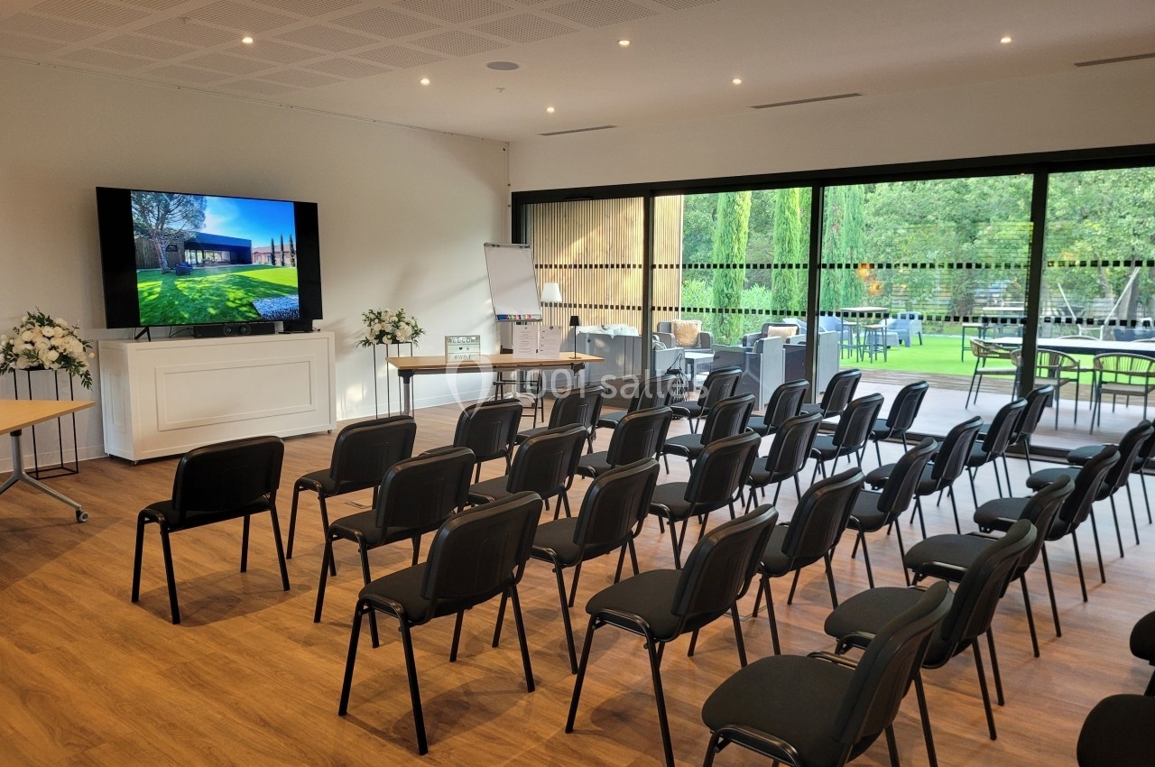 Salle de conférence lumineuse avec chaises alignées, écran allumé et vue sur un jardin à travers de grandes baies vitrées.