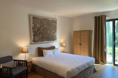 Chambre lumineuse avec lit double, fauteuil, armoire en bois, lampes de chevet et grande fenêtre donnant sur un jardin.