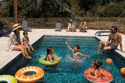 Des enfants jouent avec des bouées dans une piscine, entourés d'adultes assis sur les bords par une journée ensoleillée.
