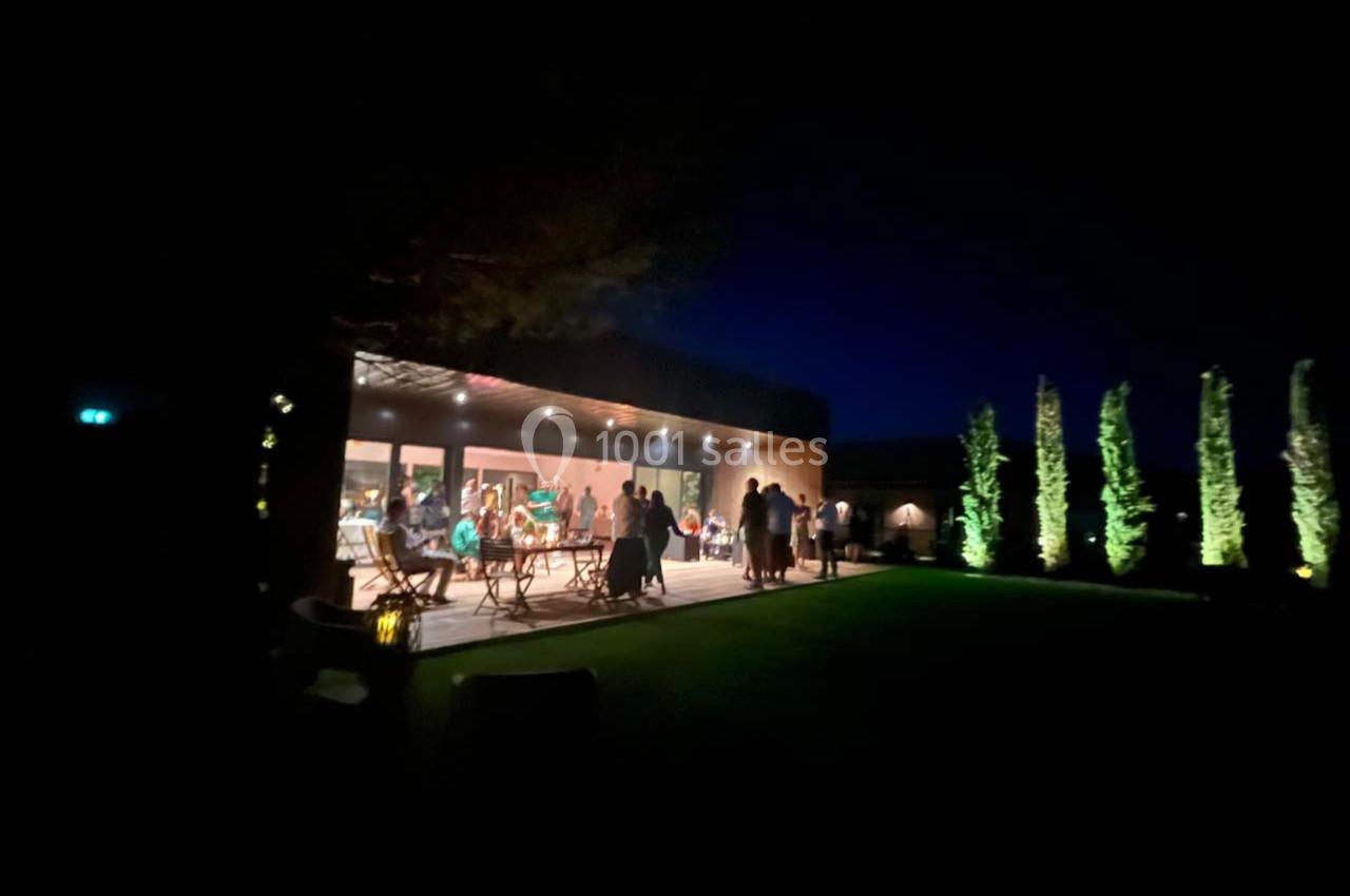 Terrasse éclairée d'une maison moderne en soirée, avec des personnes rassemblées et un jardin visible au premier plan.
