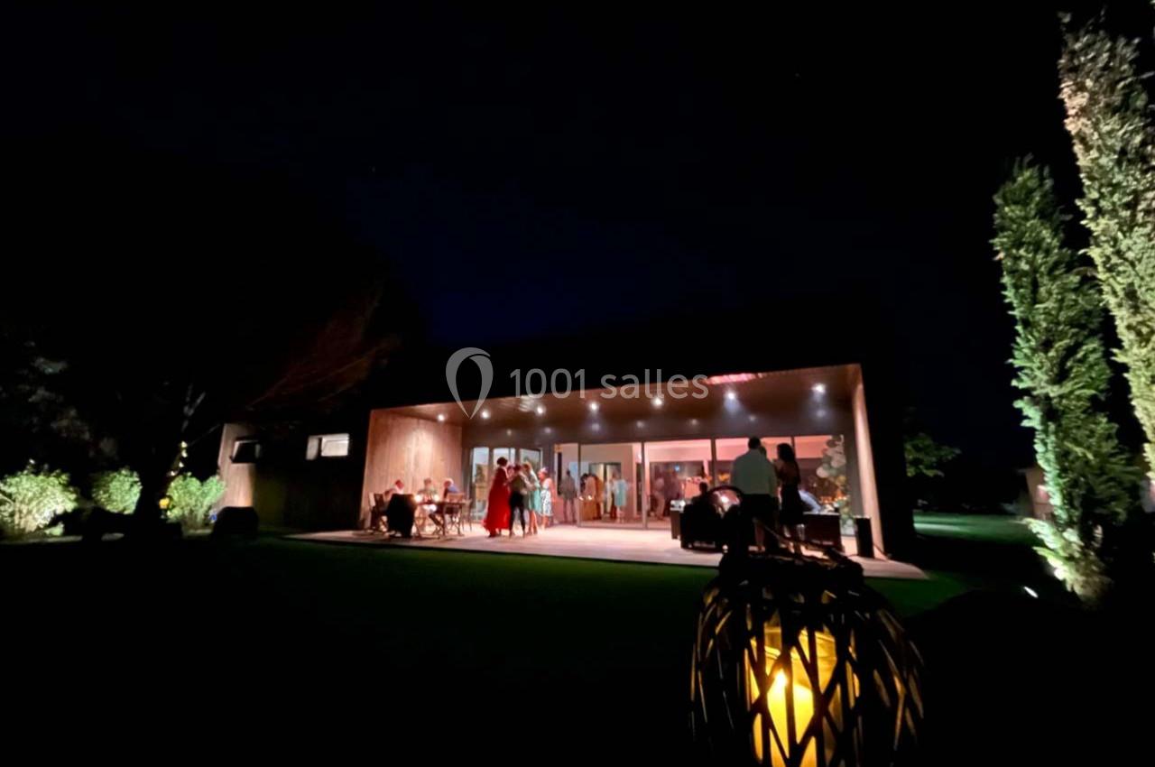 Vue nocturne d'une maison moderne éclairée, avec des personnes rassemblées sur une terrasse et un jardin en premier plan.