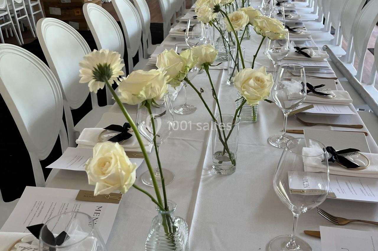 Table dressée pour un événement avec nappes blanches, fleurs blanches en vase, menus et verres alignés.