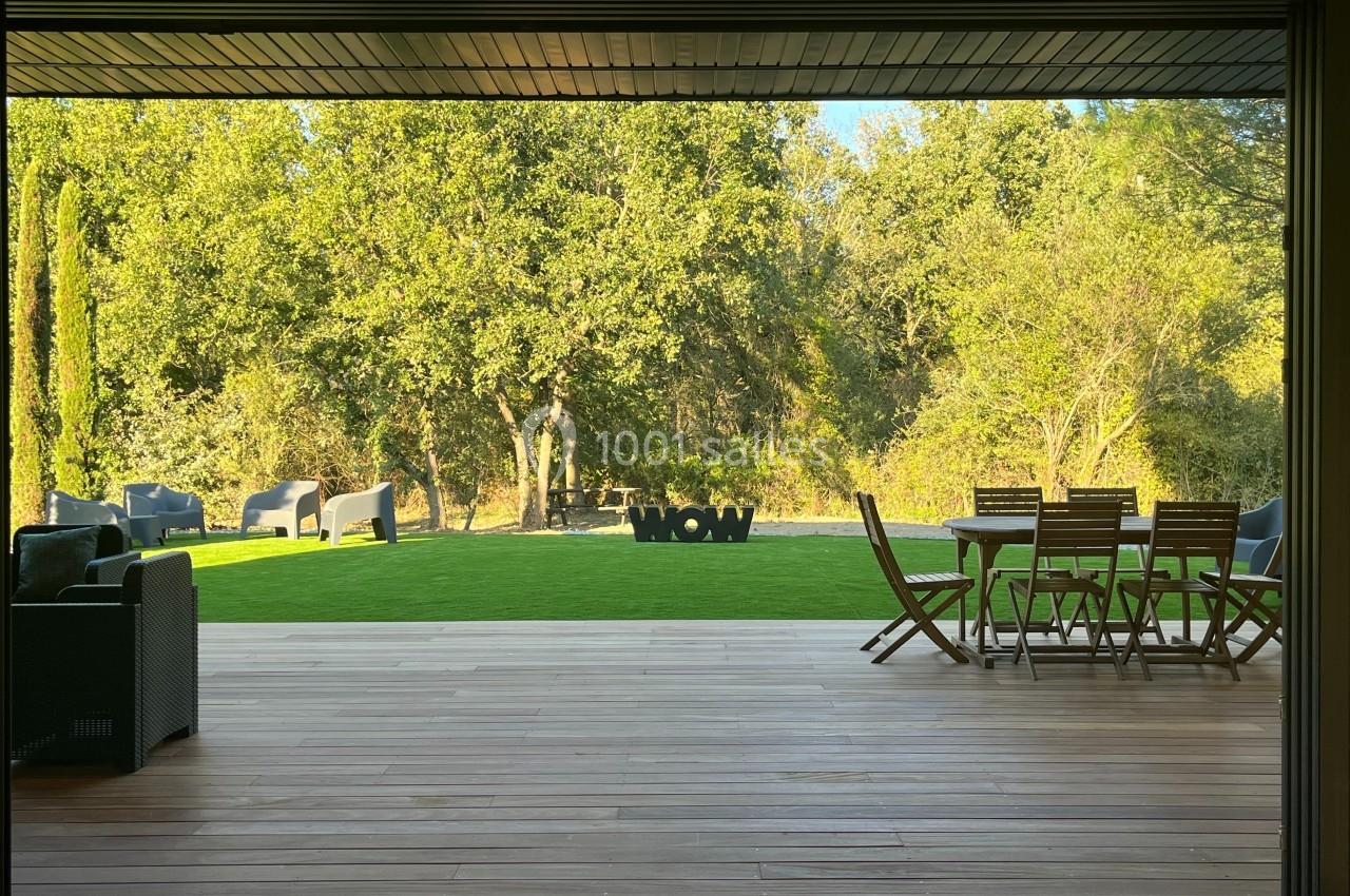 Vue sur un jardin avec pelouse, mobilier d'extérieur en bois et arbres en arrière-plan, depuis une terrasse en bois.