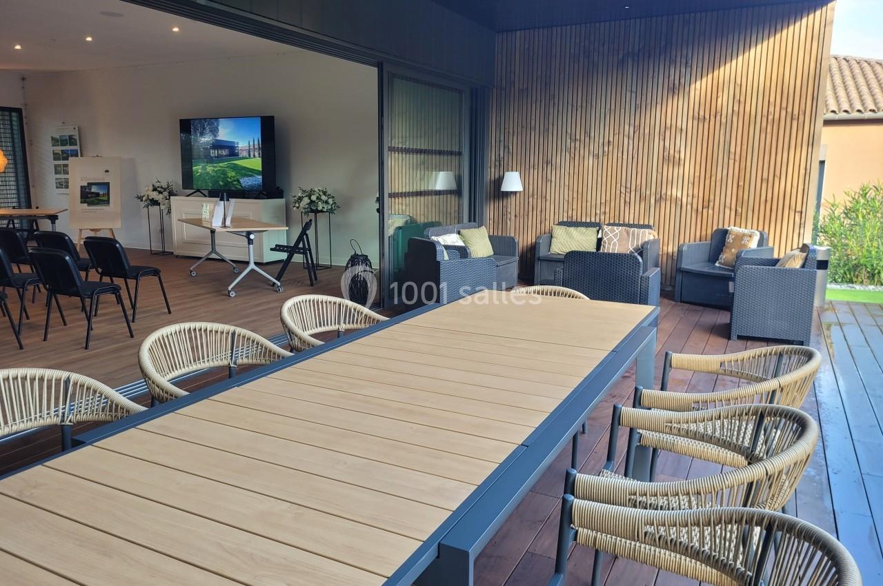 Terrasse aménagée avec une grande table en bois, des chaises en rotin et un coin salon avec fauteuils et écran en arrière…