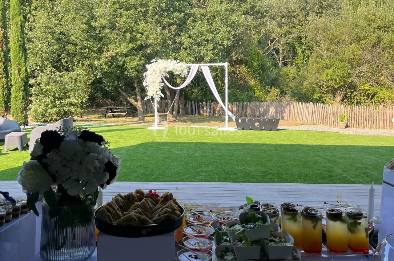 Table de réception avec boissons, desserts et fleurs en premier plan, donnant sur un jardin avec arche décorative.