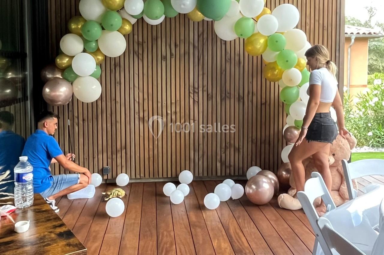 Deux personnes assemblent une arche de ballons colorés sur une terrasse en bois, près d'un mur en lambris.